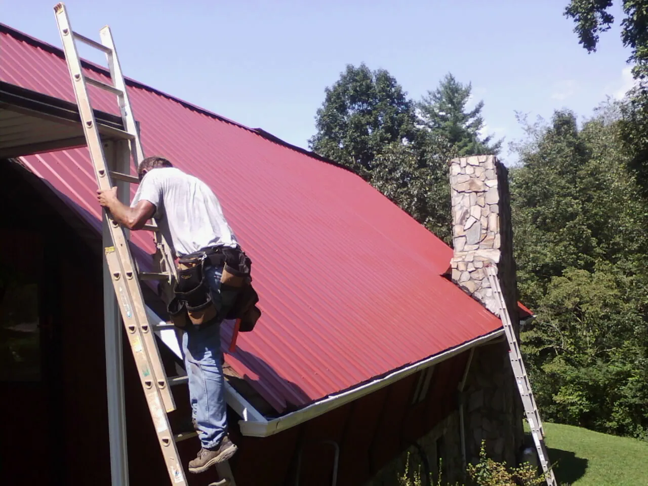 Roofer on a ladder beside a red metal roof and stone chimney — Soffit & Fascia Repair by Roofing Brothers in Merrittstown